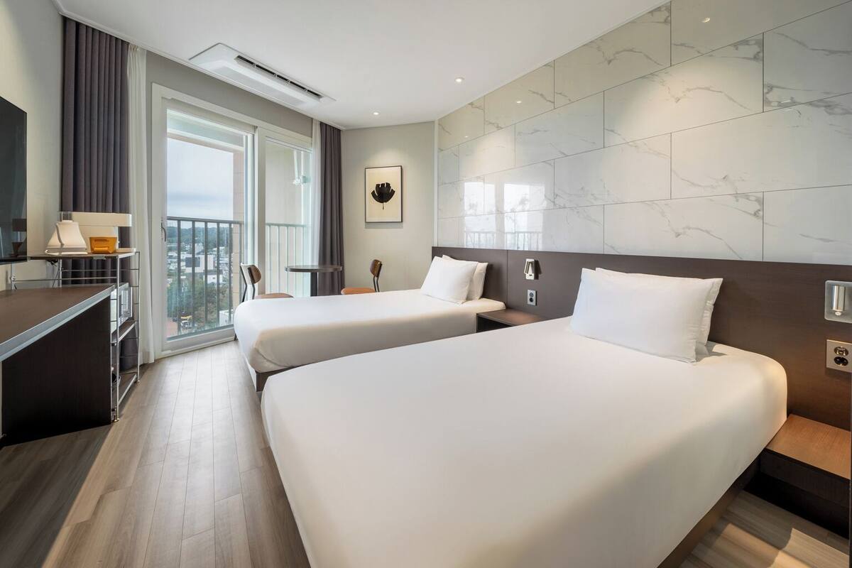 Ein Hotelzimmer mit zwei Betten, einem Schreibtisch, einem Sessel, einem kleinen Tisch und einem großen Fenster mit Blick.