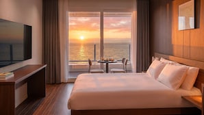 Une chambre d’hôtel avec un lit, une télévision, une table à manger avec des chaises et une vue sur l’océan au coucher du soleil.