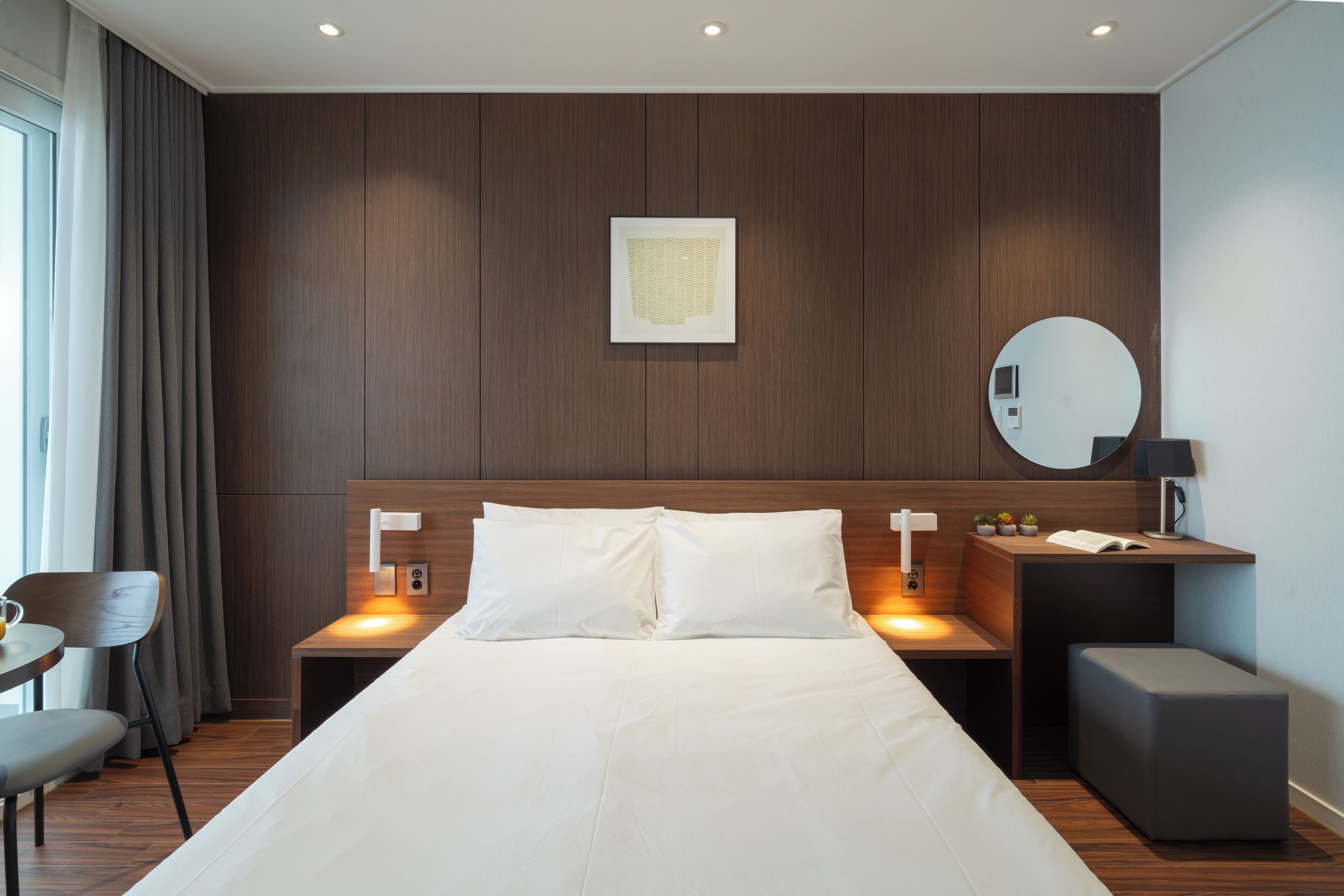 Una habitación de hotel moderna con una cama grande, dos mesitas de noche, un espejo redondo y una pequeña mesa con una lámpara.