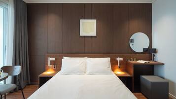 Una habitación de hotel moderna con una cama grande, dos mesitas de noche, un espejo redondo y una pequeña mesa con una lámpara.