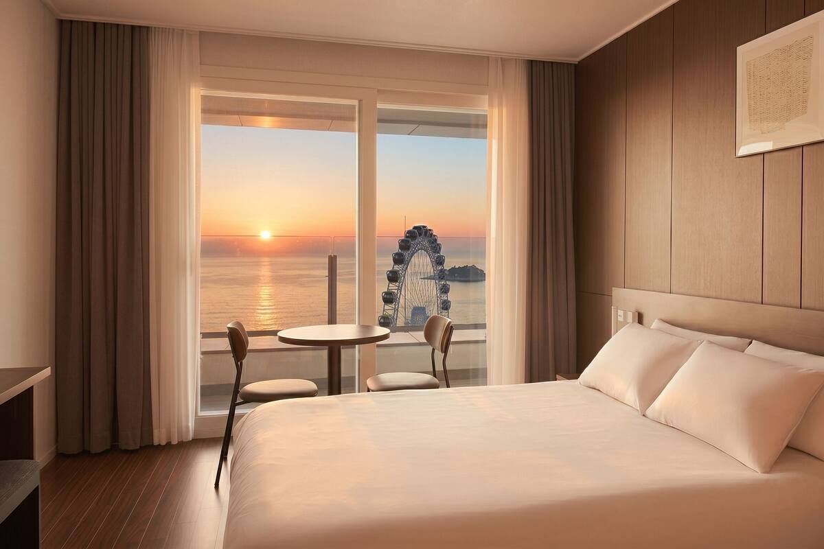Ein Hotelzimmer mit einem großen Bett, einem kleinen Tisch, zwei Stühlen und Blick auf einen Sonnenuntergang sowie ein Riesenrad.