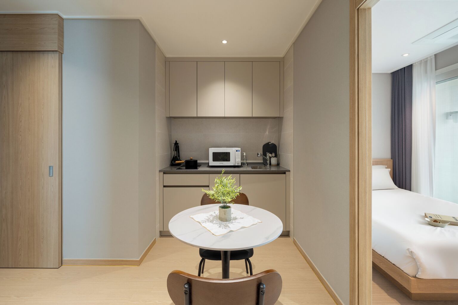 A modern hotel room with a compact kitchenette, a round dining table with a vase, and a bed with a window view.