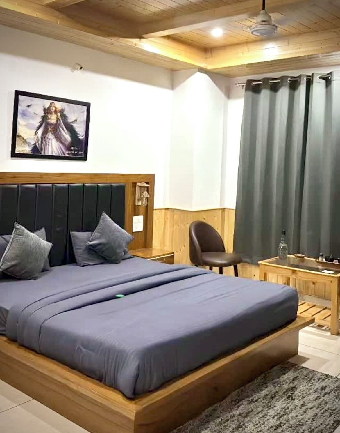 Un dormitorio con una cama de madera, un escritorio, una silla, un ventanal con cortinas y un cuadro en la pared.