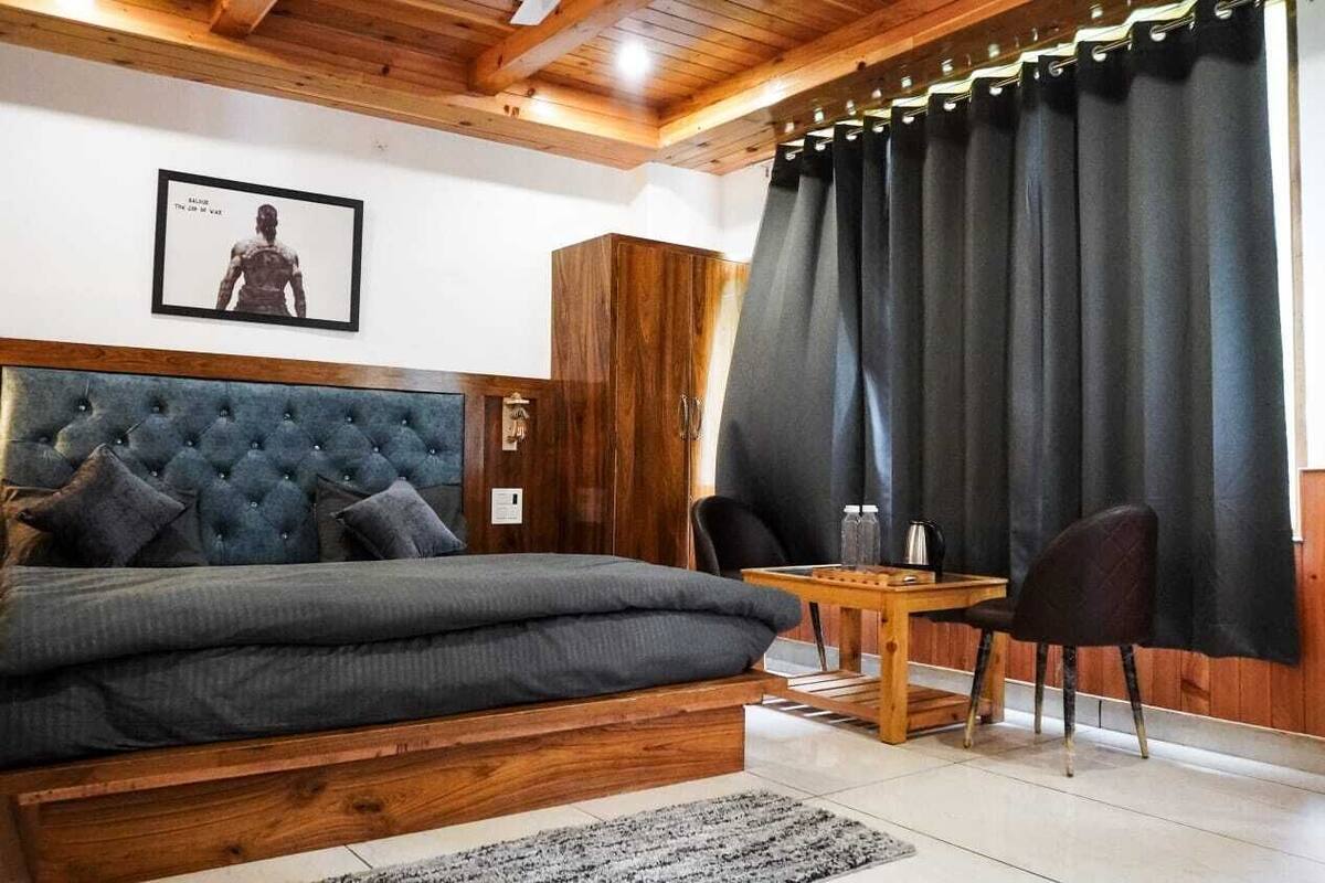 A bedroom with a large bed, a wooden headboard, a small table with chairs, and a dark curtain.