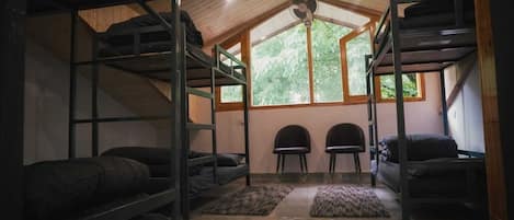 Un dormitorio con literas, un ventilador, dos sillas y una ventana con vistas a árboles.