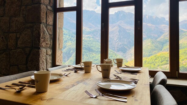 A dining table set with plates, cups, and cutlery, overlooking a mountainous landscape through large windows. - Askireg Hotel (Pülümür)