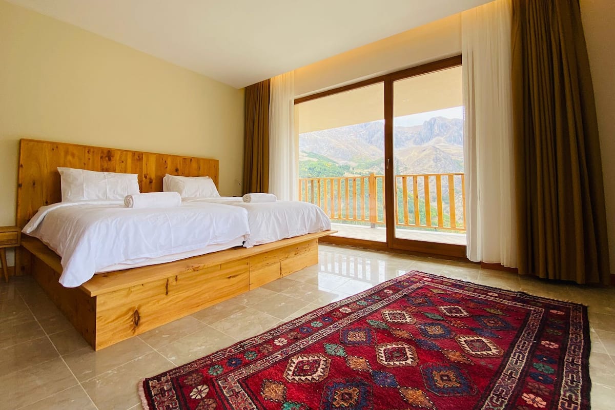 A double bed with a wooden headboard, a large window with curtains, and a view of mountains.