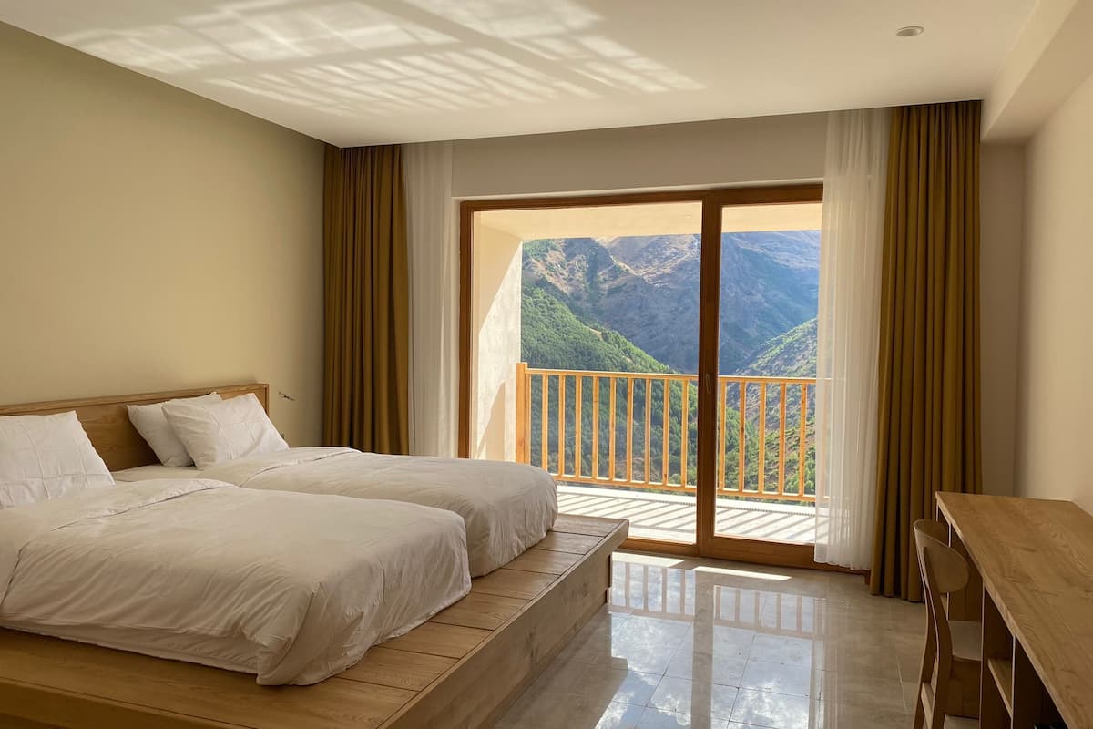 A hotel room with a large bed, a wooden desk, and a balcony with a mountain view.