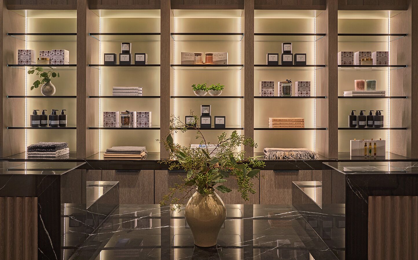 A modern bathroom with marble countertops, a central vase with greenery, and shelves with toiletries.