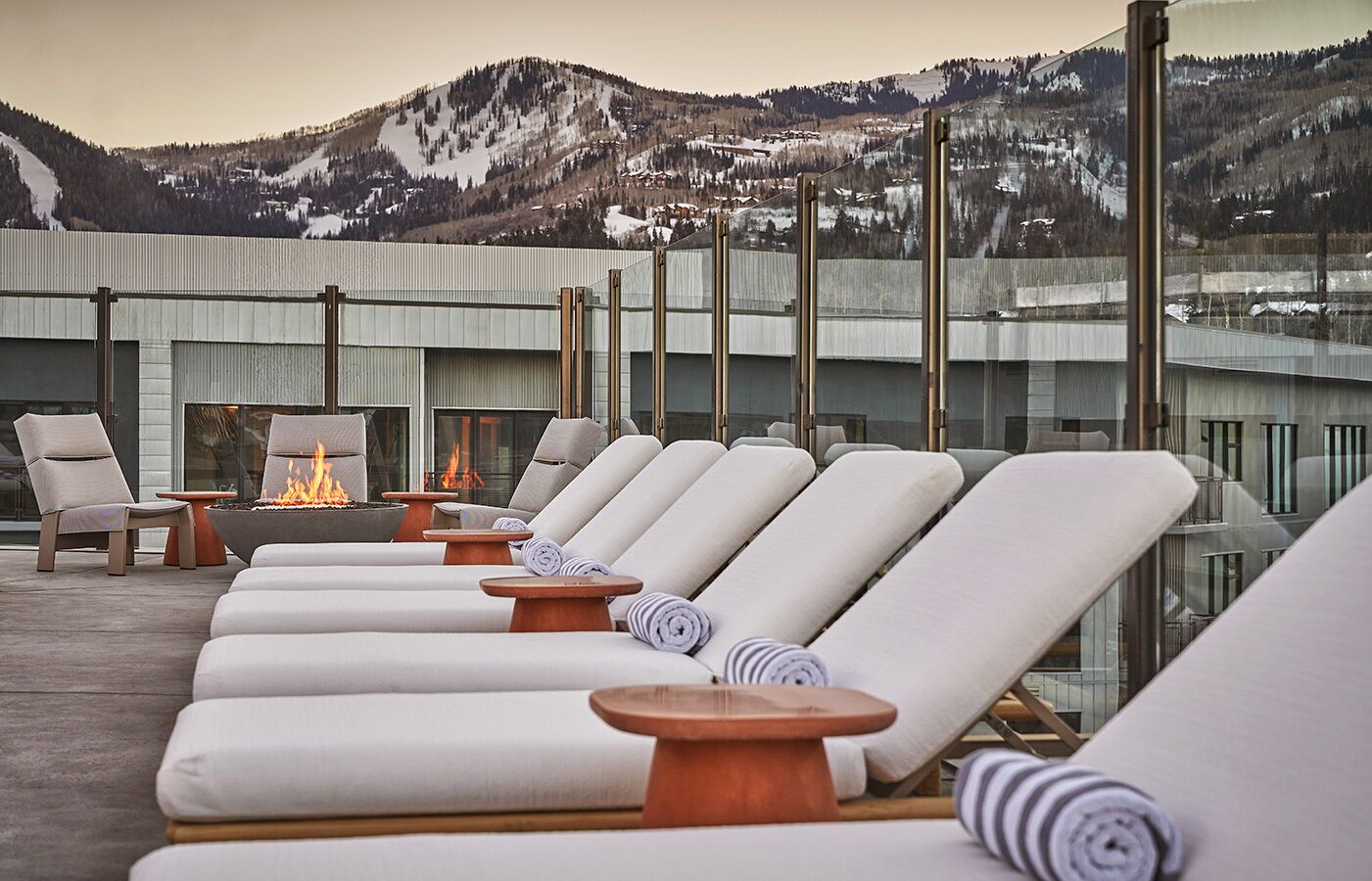 A rooftop lounge area with sun loungers, a fire pit, and a view of snow-covered mountains.