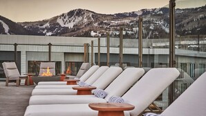 A rooftop lounge area with sun loungers, a fire pit, and a view of snow-covered mountains.