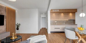 A modern living room with a flat-screen TV, a black coffee table, and a sofa. There is a wooden slat wall and a potted plant. The kitchen has wooden cabinets, a sink, and a dining area with a glass table and chairs. - 03. Luxury Apartments L&L in Tucepi (Tucepi)