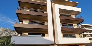 A multi-story building with balconies, a flat roof, and a mountainous backdrop. - 03. Luxury Apartments L&L in Tucepi (Tucepi)
