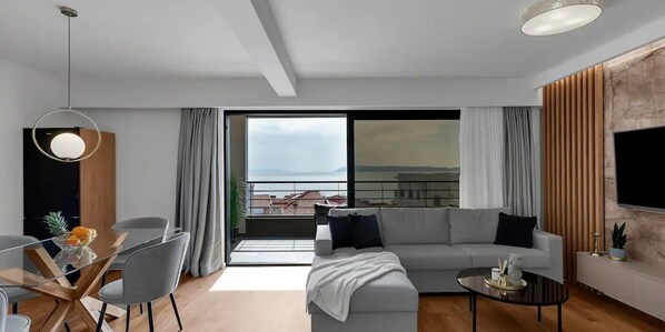 A modern living room with a grey sofa, a glass dining table, and a black coffee table. There is a large window with a view of buildings and a balcony. - 03. Luxury Apartments L&L in Tucepi (Tucepi)