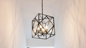 Eine geometrische Pendelleuchte mit drei Glühbirnen, die an einer Kette hängen und von der Decke in einem Raum mit weißen Wänden und einem Fenster im Hintergrund befestigt sind.