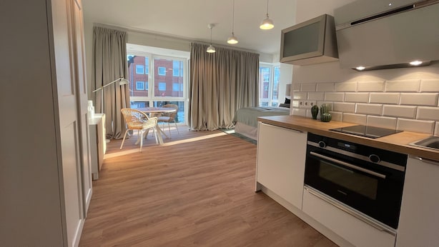 Una cocina moderna con horno empotrado, un comedor con mesa y sillas, y vistas a un edificio de ladrillo a través de grandes ventanales.