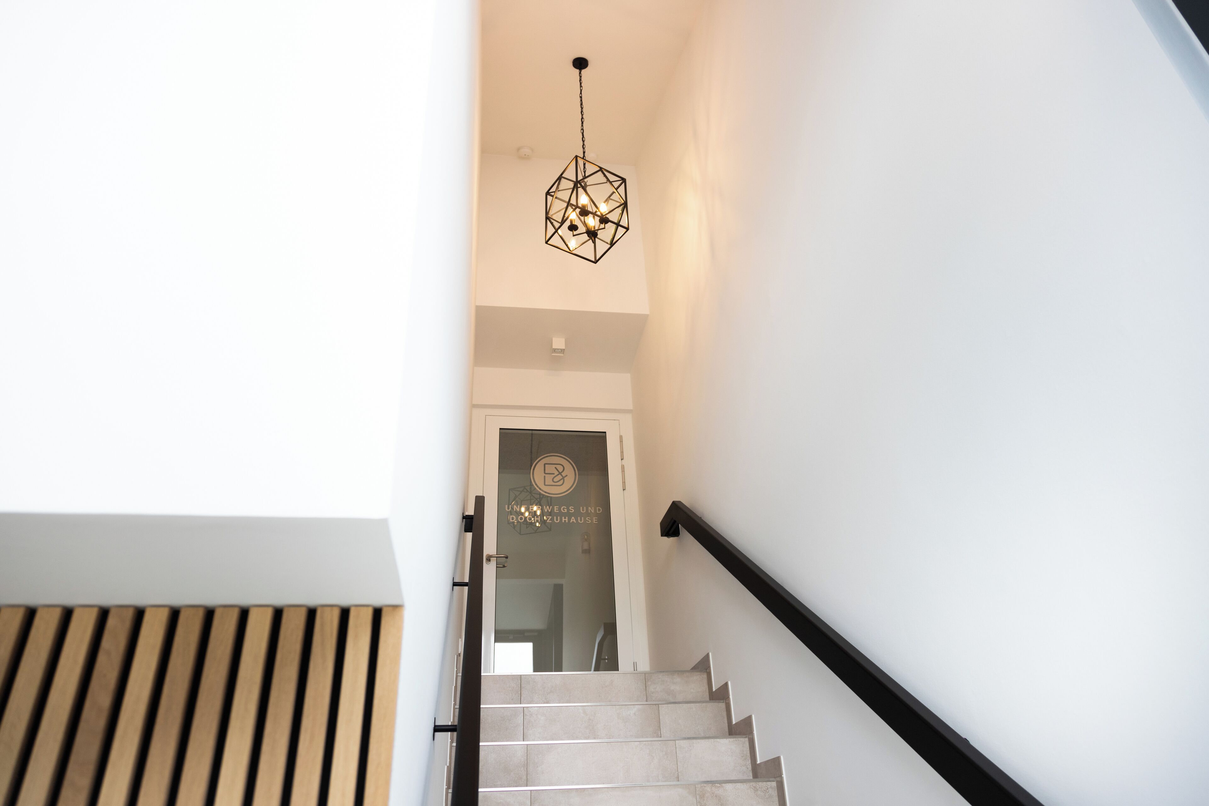 Eine Treppe mit einer geometrischen Pendelleuchte, einer Wand mit Holzlamellen und einer Tür mit einem Logo.