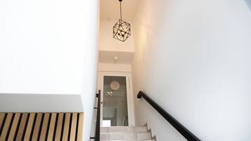 Eine Treppe mit einer geometrischen Pendelleuchte, einer Wand mit Holzlamellen und einer Tür mit einem Logo.