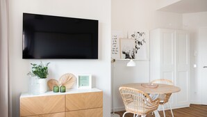 A modern living room with a flat-screen TV mounted on the wall, a wooden cabinet with decorative items, a round dining table with wicker chairs, and a white door in the background.