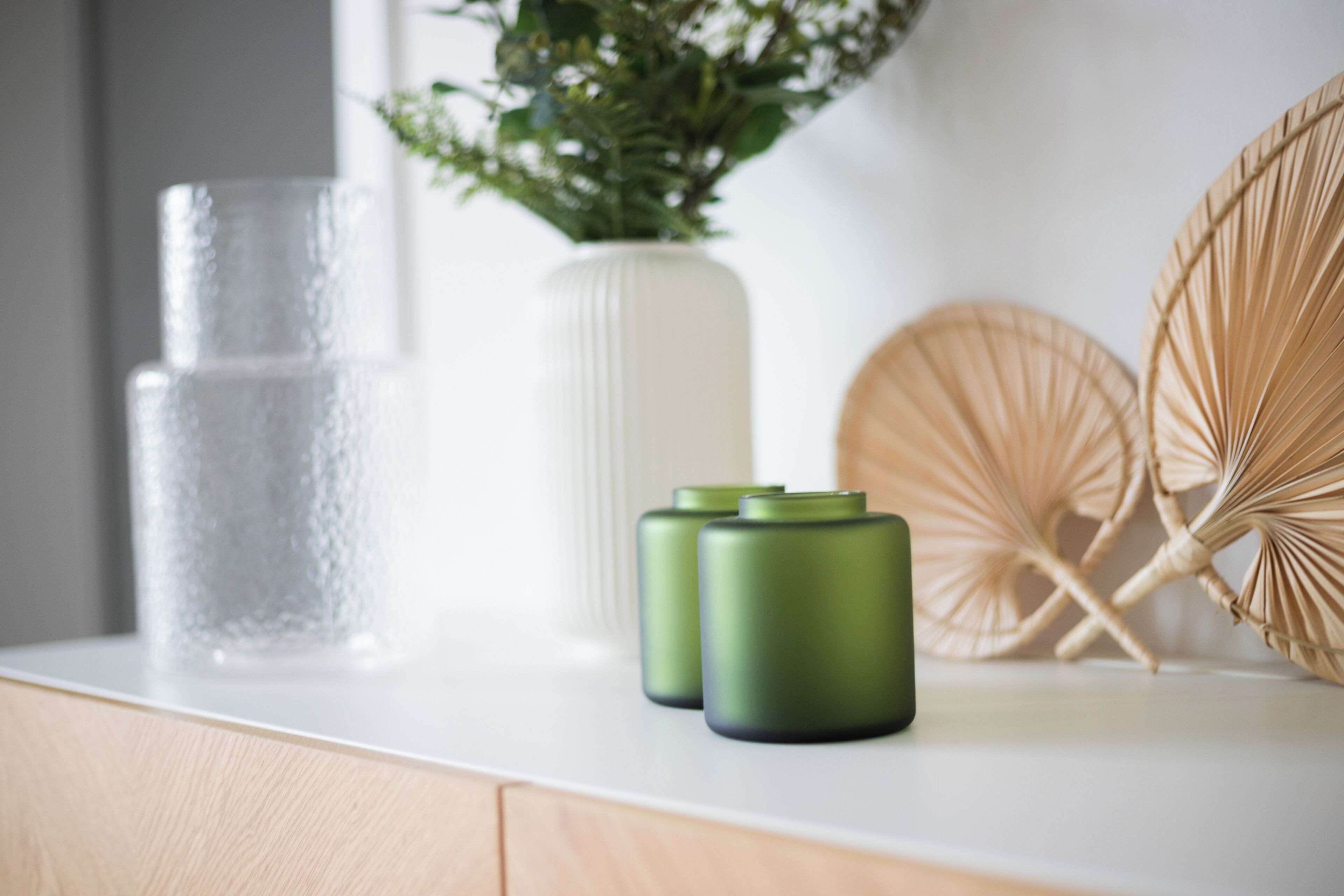 Eine weiße Arbeitsplatte mit einem klaren Glas, einer weißen Vase mit Grünpflanzen und zwei grünen zylindrischen Objekten.