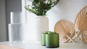 A white countertop with a clear glass, a white vase with green foliage, and two green cylindrical objects.