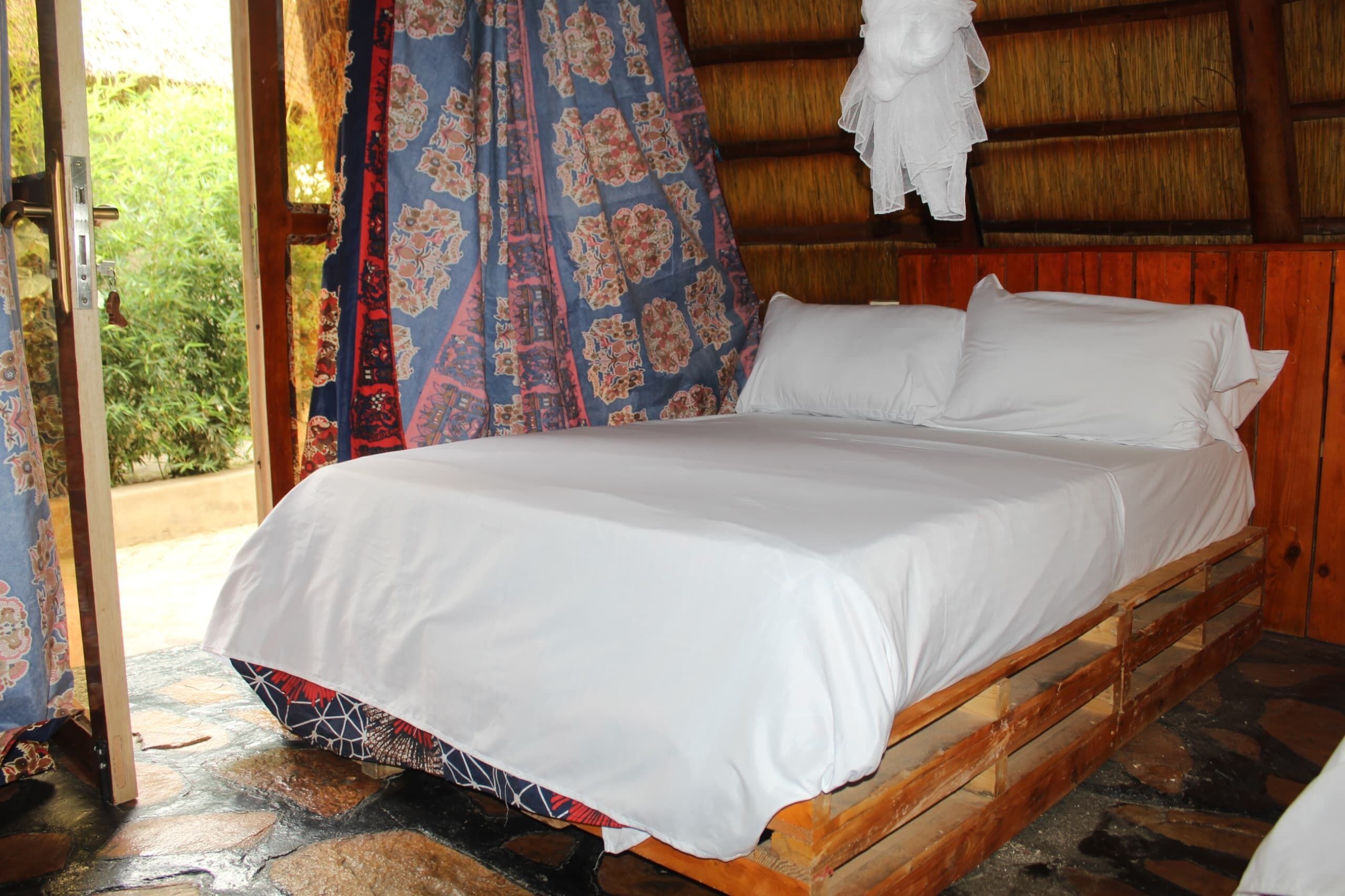 A bed in a rustic room with a wooden headboard, a patterned curtain, and a view of greenery outside.