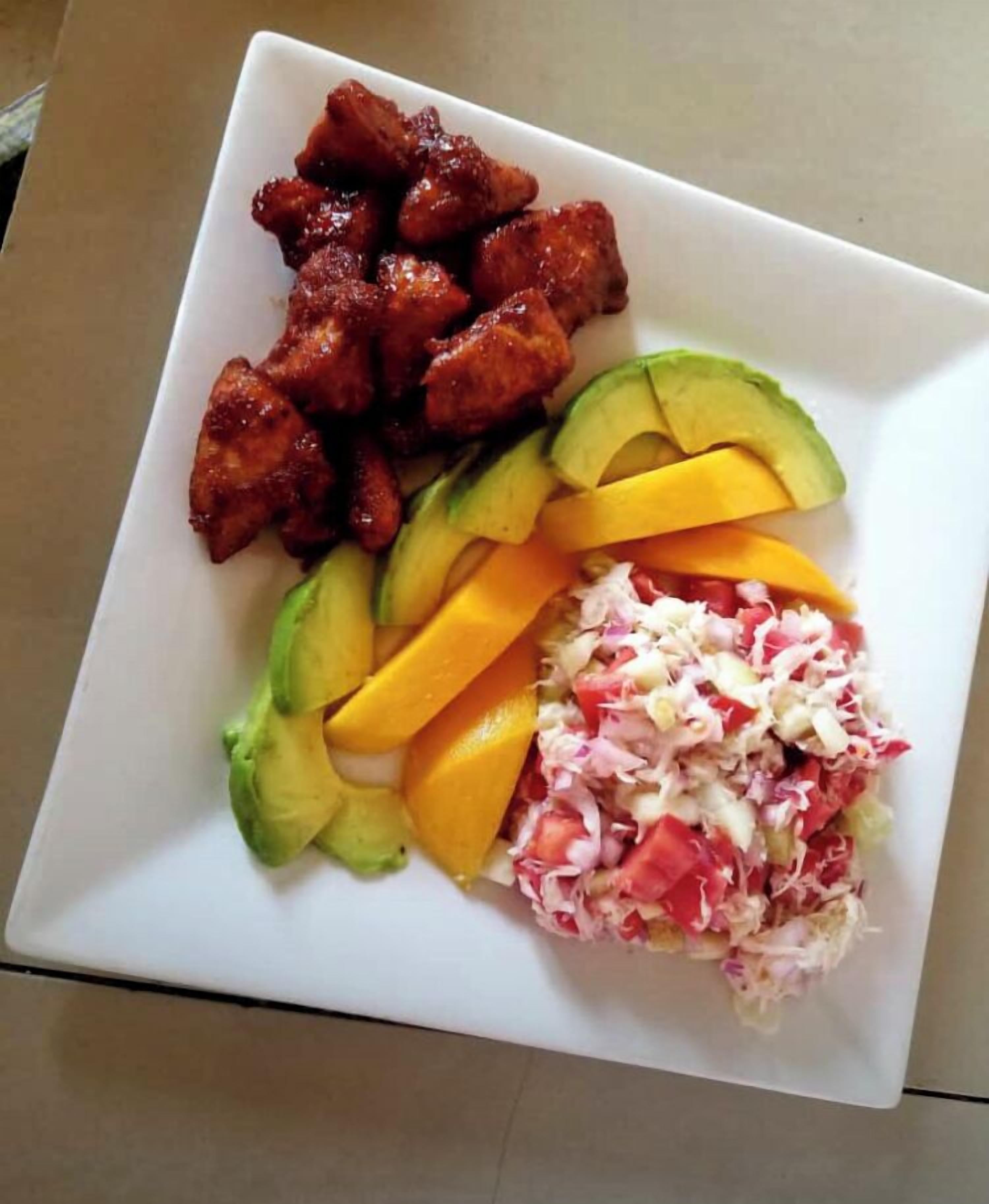 Een bord met gegrilde kip, plakjes avocado, mango en een koolsalade.