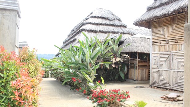 A pathway lined with tropical plants and red flowers, leading to thatched-roof huts with wooden walls.