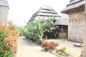 Un sentier bordé de plantes tropicales et de fleurs rouges, qui mène à des huttes aux toits de chaume et aux murs en bois.