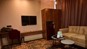Une chambre d’hôtel comprenant une armoire en bois, un canapé, un bureau avec une chaise, une télévision, une petite table avec une bouilloire et une poubelle.