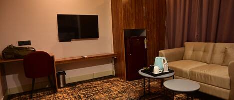 A hotel room with a wooden wardrobe, a sofa, a desk with a chair, a television, a small table with a kettle, and a trash bin.
