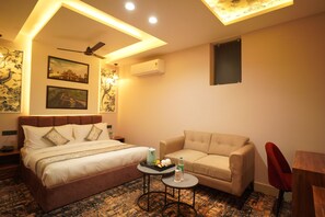 A hotel room with a bed, a sofa, a small table with a tray of fruit and a bottle of water, a ceiling fan, and wall-mounted air conditioning.