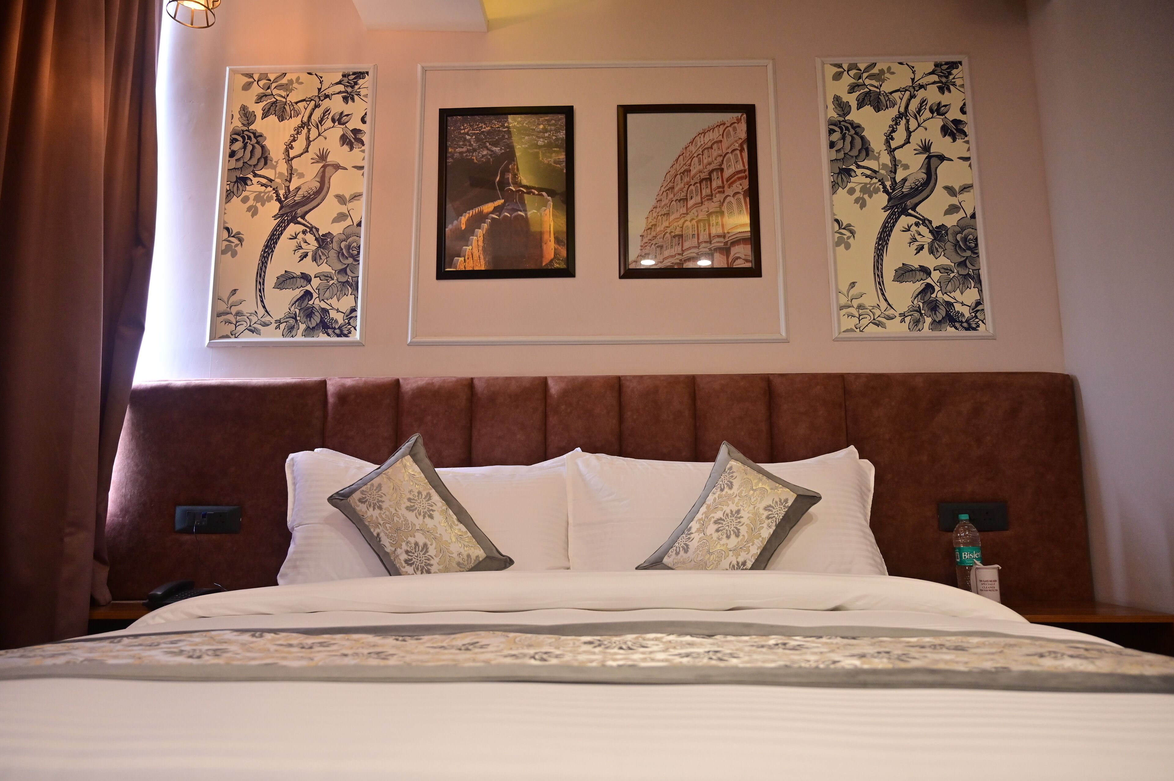 A hotel room with a large bed, two framed artworks above the bed, a brown headboard, and a nightstand with a water bottle.