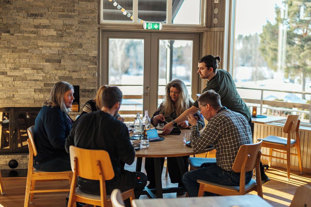 A group of people seated around a wooden table in a room with a brick wall and large windows.