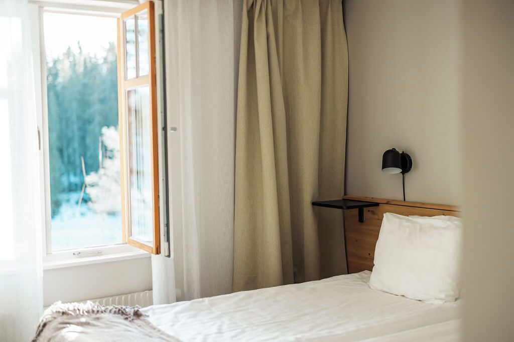 A bedroom with a bed, a nightstand, a lamp, a window with curtains, and a view of trees outside.