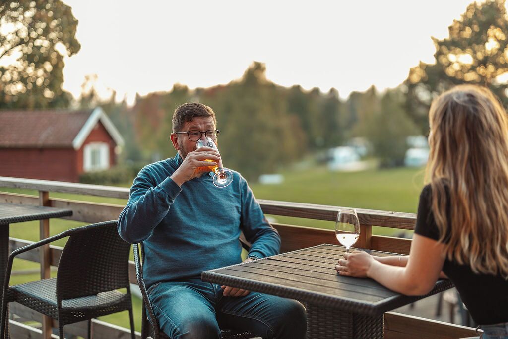Två personer sitter vid ett bord utomhus, och på bordet står ett glas vin, ett glas öl och en ölflaska.
