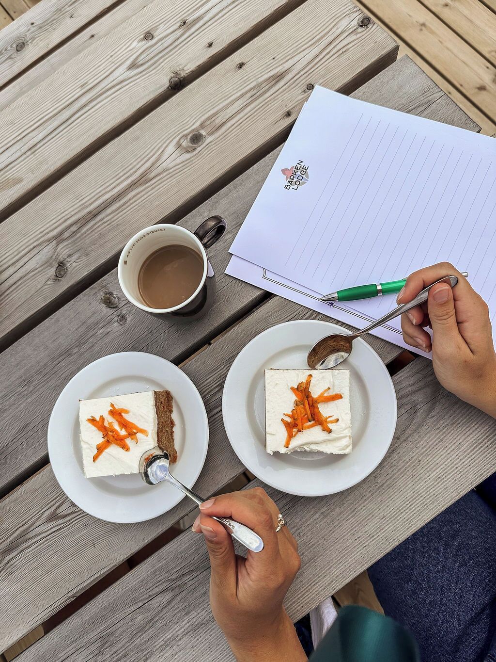 A wooden table with a notebook, a pen, a cup of coffee, and two plates of cake with carrot garnish.
