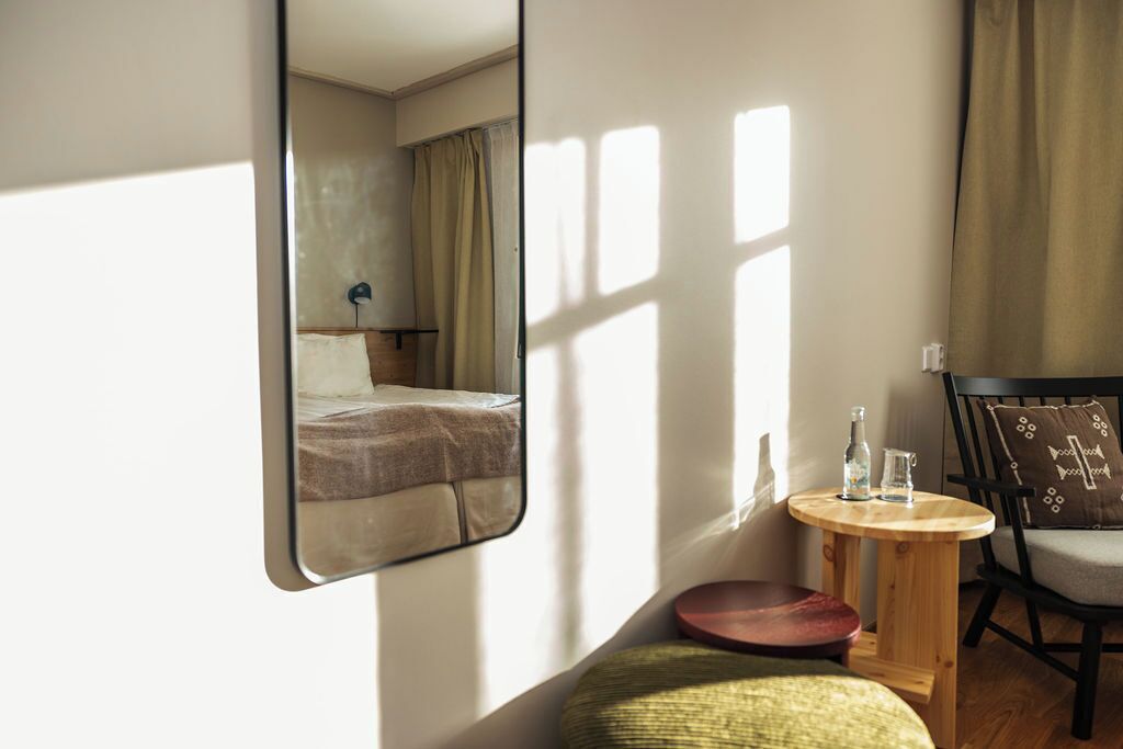 A hotel room with a bed, a round wooden table with a glass bottle and a glass, a green cushion, and a chair with a patterned cushion.