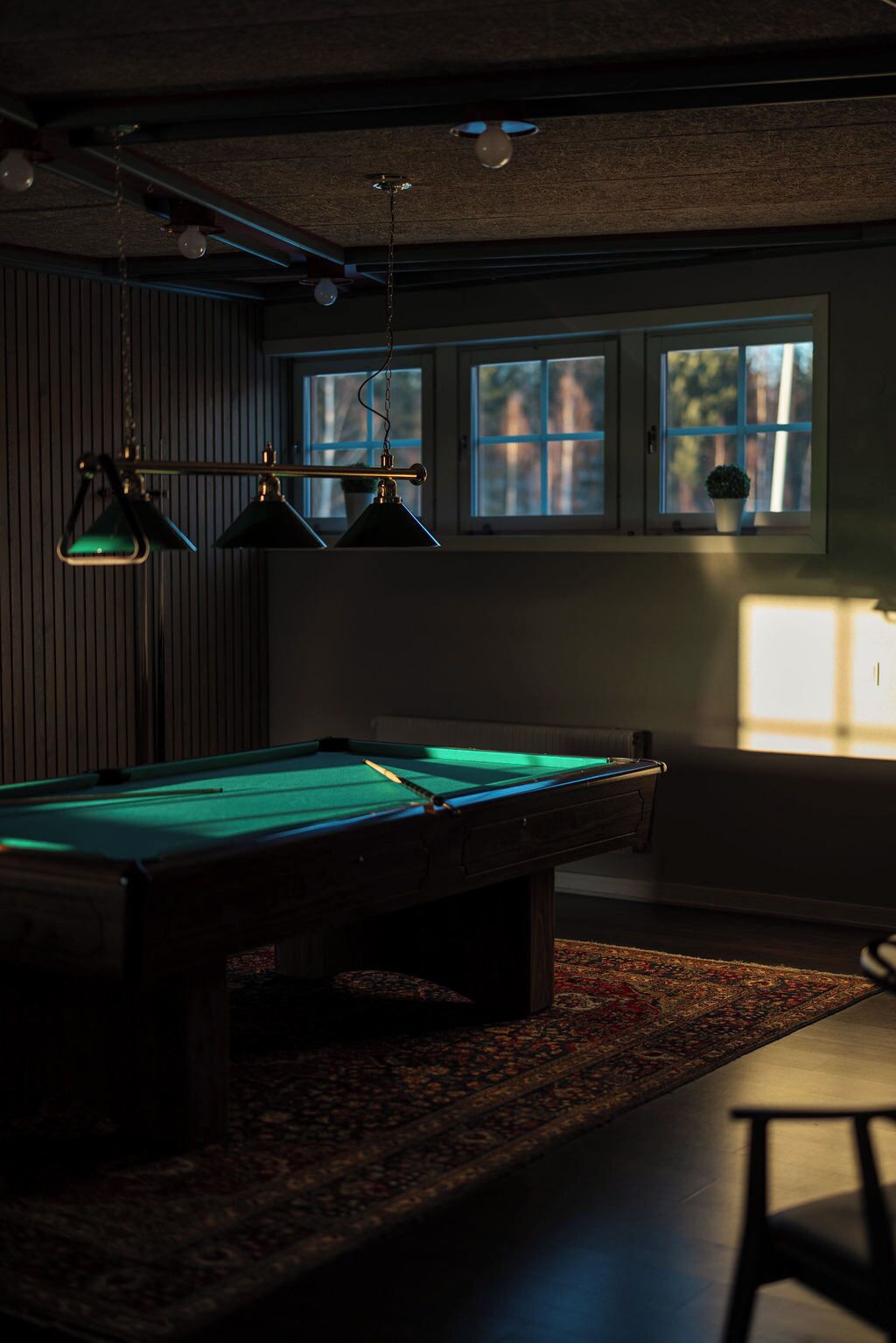 Una mesa de billar iluminada desde arriba, un tapete estampado y una ventana con persianas.