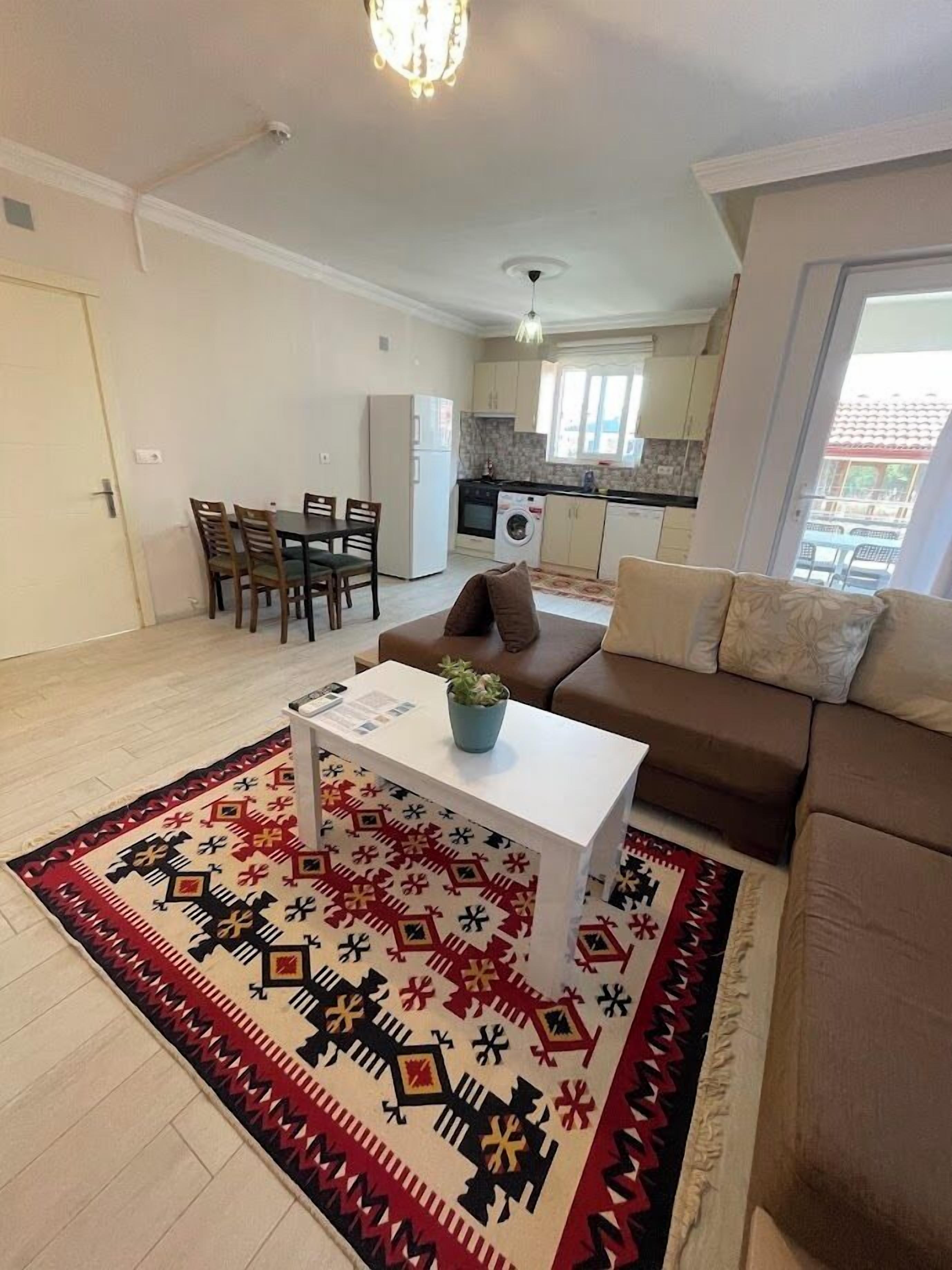 Een woonkamer met een bank, salontafel en een rug met patroon. Er is een eethoek met stoelen en een tafel, en een keuken op de achtergrond.