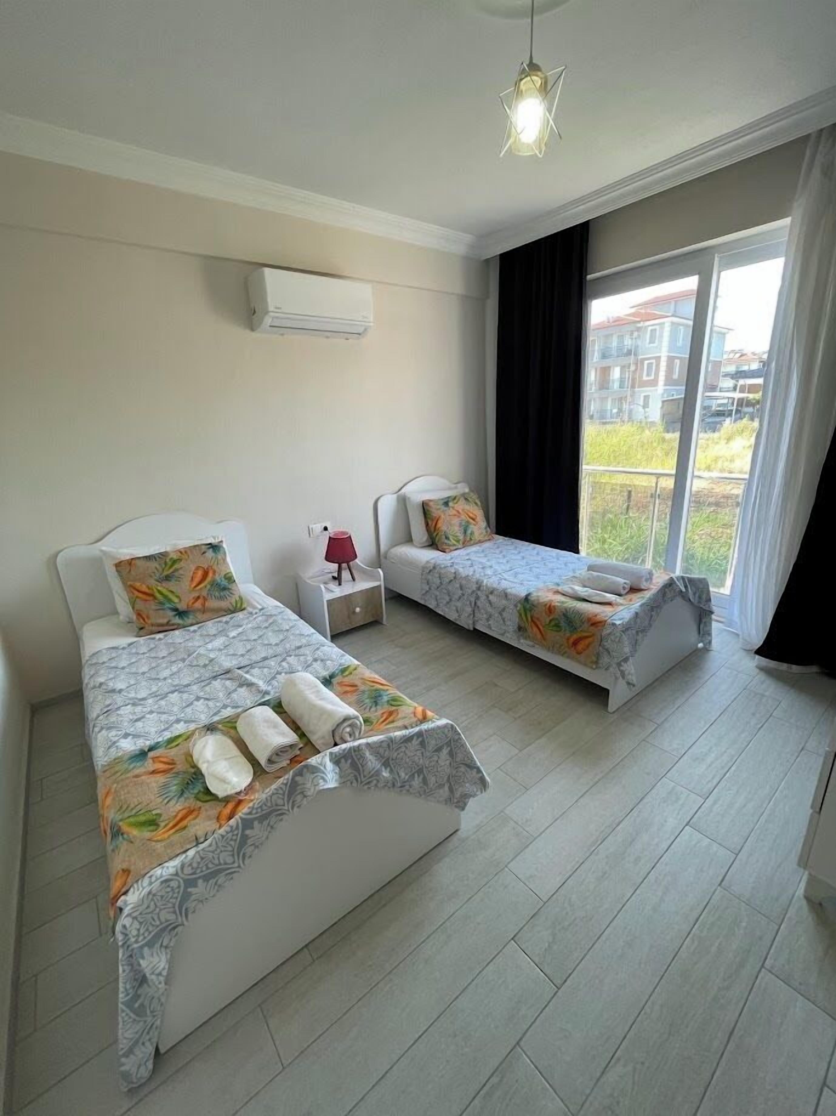 Une chambre d’hôtel avec deux lits, une petite table, une lampe rouge et une vue sur les immeubles par la fenêtre.