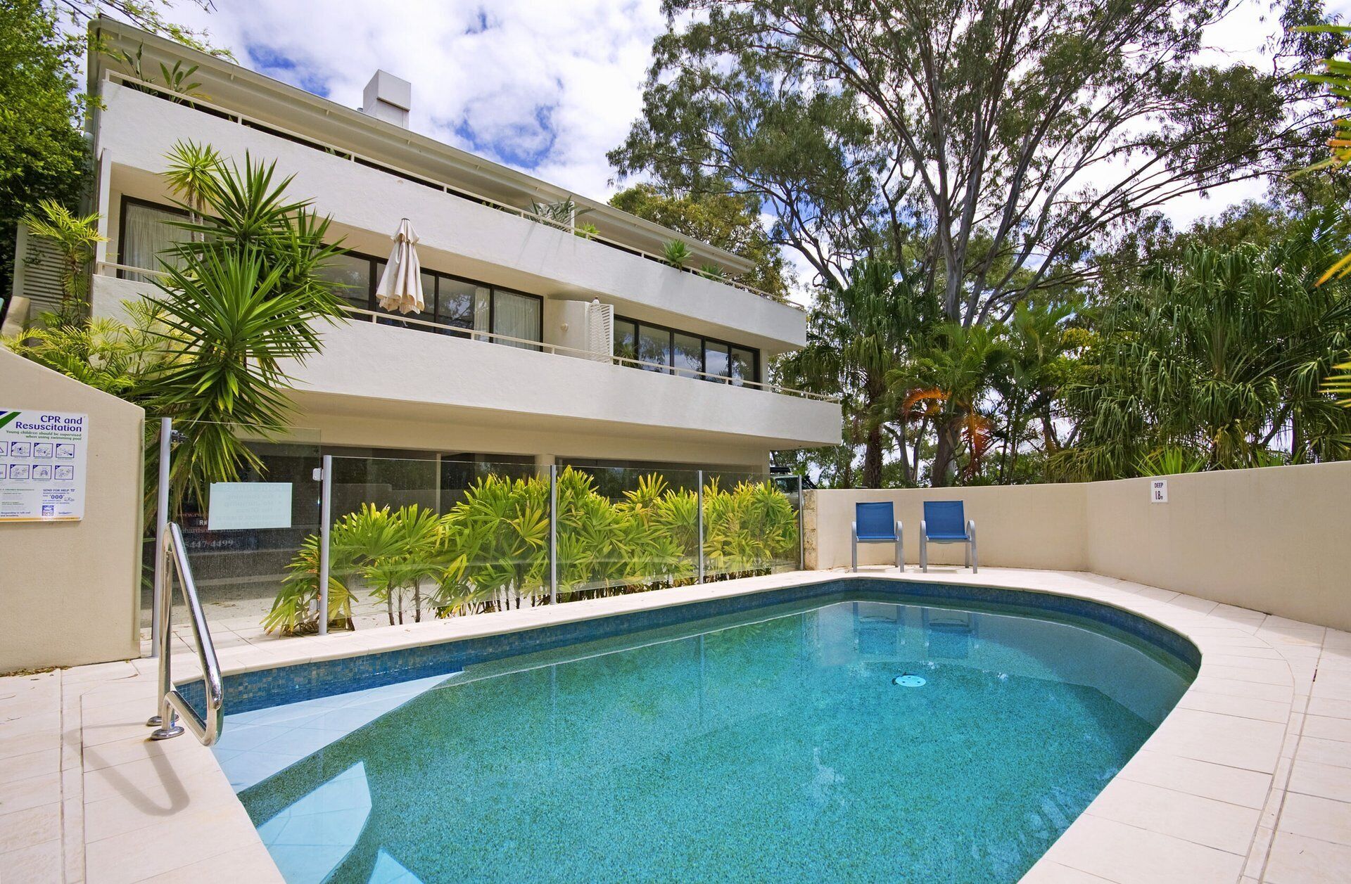 A swimming pool with a clear blue water, surrounded by a tiled deck, and a two-story building with balconies in the background.