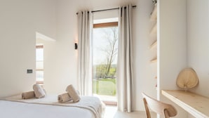 A bedroom with a large bed, a wooden chair, a wooden shelf, and a view of the outdoors through a window.