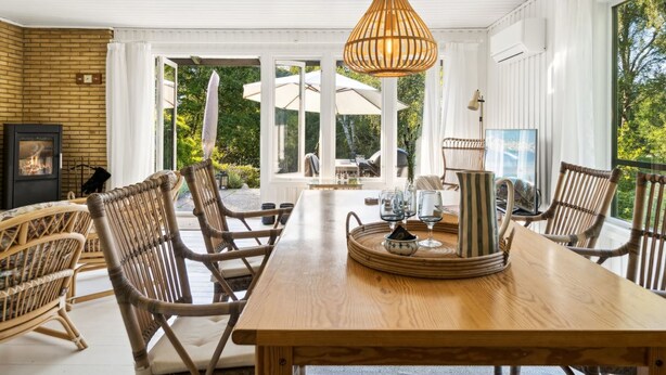 Una mesa de comedor de madera con sillas de mimbre, una lámpara colgante y vistas al exterior a través de amplios ventanales.
