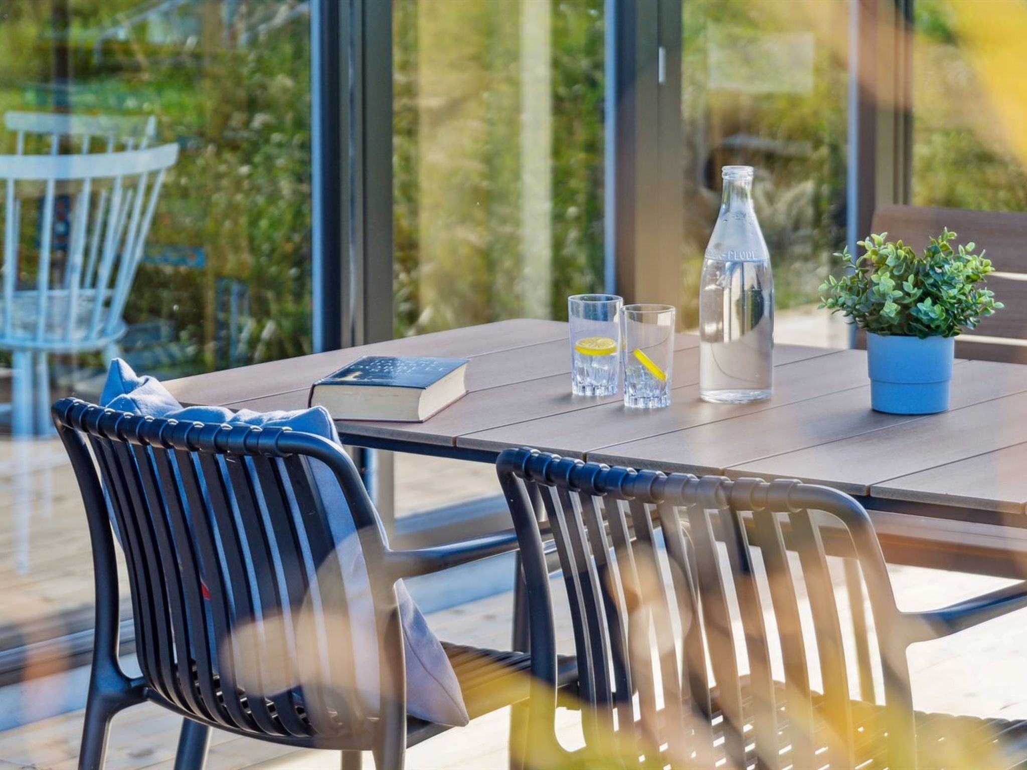 A modern outdoor dining area with a table, chairs, a book, glasses, a water bottle, and a potted plant.