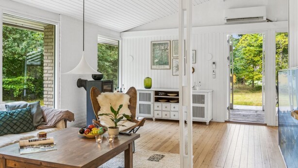 A modern, well-lit room with a wooden floor, a dining table with a bowl of fruit, a sofa with cushions, and a large window offering a view of greenery outside.