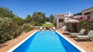 Una piscina rectangular con borde de azulejos, rodeada de sillas de descanso y sombrillas, con un edificio blanco de techo y pared de piedra de fondo.
