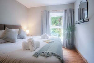 A neatly made bed with pillows, a blanket, and towels, a bedside lamp, a window with curtains, and a potted plant.