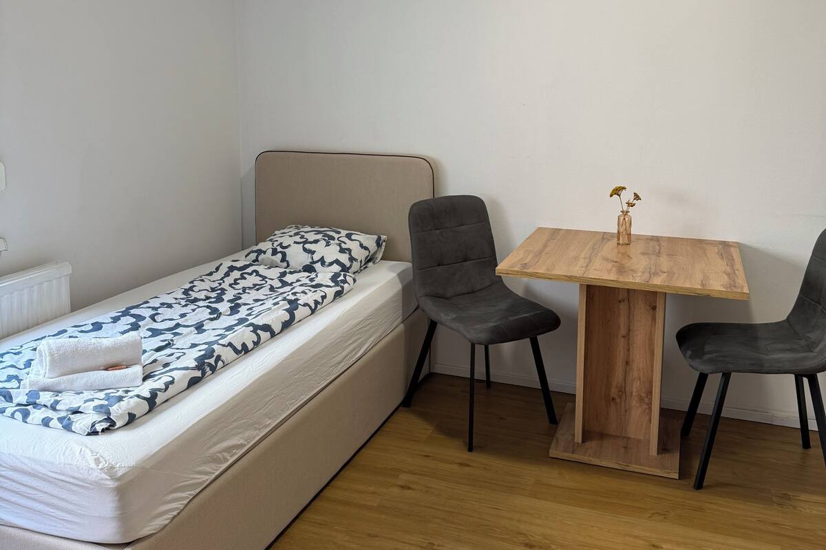 Una cama individual con una manta estampada, una mesita de madera con dos sillas y un radiador.