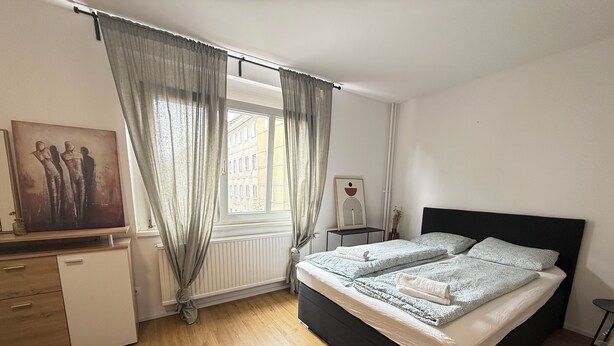 Een slaapkamer met een bed, een dressoir, een raam met gordijnen en een schilderij aan de muur.